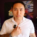 Christopher To, a business coach, wearing a white patterned shirt with a clip-on microphone, posing confidently indoors against a colorful, abstract backdrop.