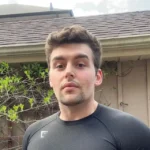 Nic Paul Anthony, an Online Fitness Coach, standing outdoors in a black athletic shirt, with a neutral expression against a backdrop of greenery and a house roofline.