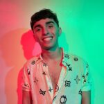 Jayden Adamson, an Amazon FBA Coach, smiling confidently while wearing a patterned shirt, standing against a vibrant backdrop illuminated with red and green lighting.
