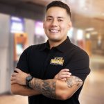 A well-known American entrepreneur, Paul Alex, founder of 8-figure businesses like ATM Together, smiling confidently in a black polo shirt with the company logo, standing against a blurred indoor background featuring ATMs.